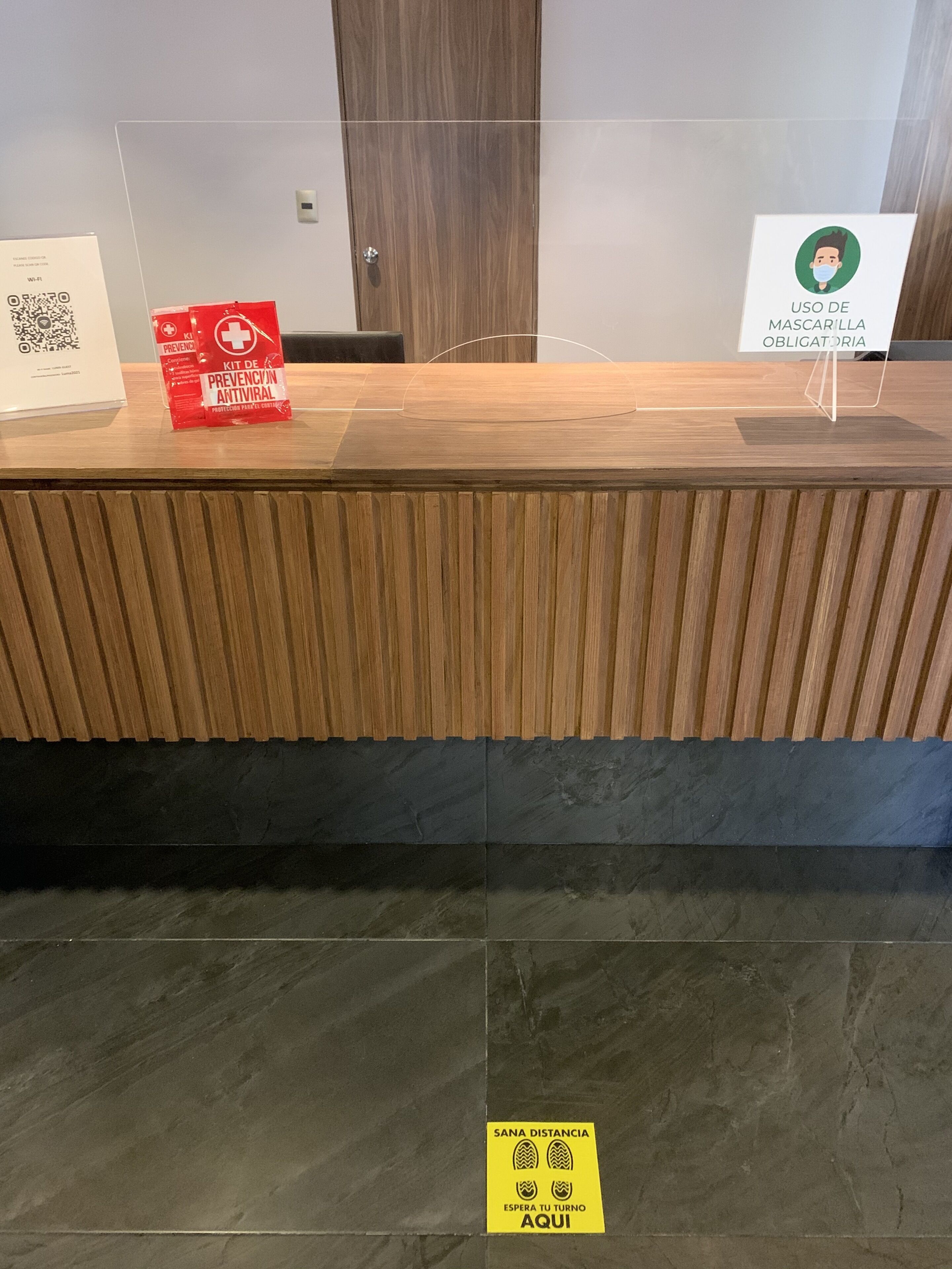 Een receptiebalie met een QR-code, een rode preventiekit voor COVID-19 en een bordje dat verplichting tot het dragen van een mondmasker aangeeft.