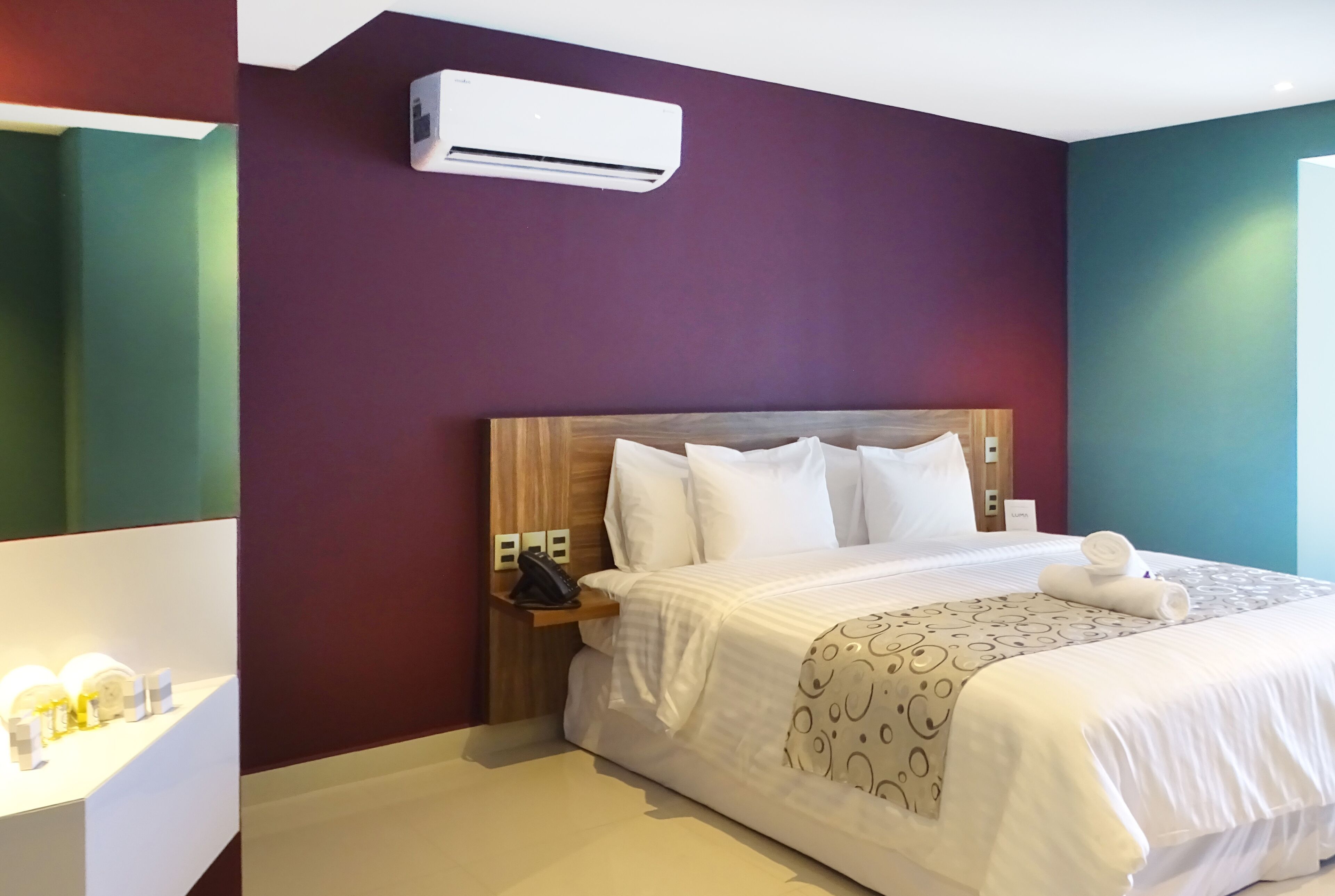 A hotel room with a wooden headboard, white bedding, a bedside table with a lamp, and an air conditioning unit.