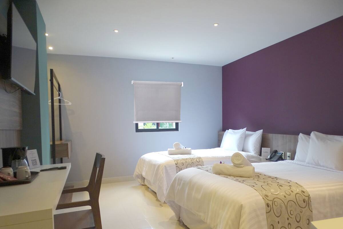 Une chambre d’hôtel avec deux lits, un bureau et une chaise. Il y a une fenêtre avec un store blanc, un téléphone sur la table de chevet et une serviette sur chaque lit.