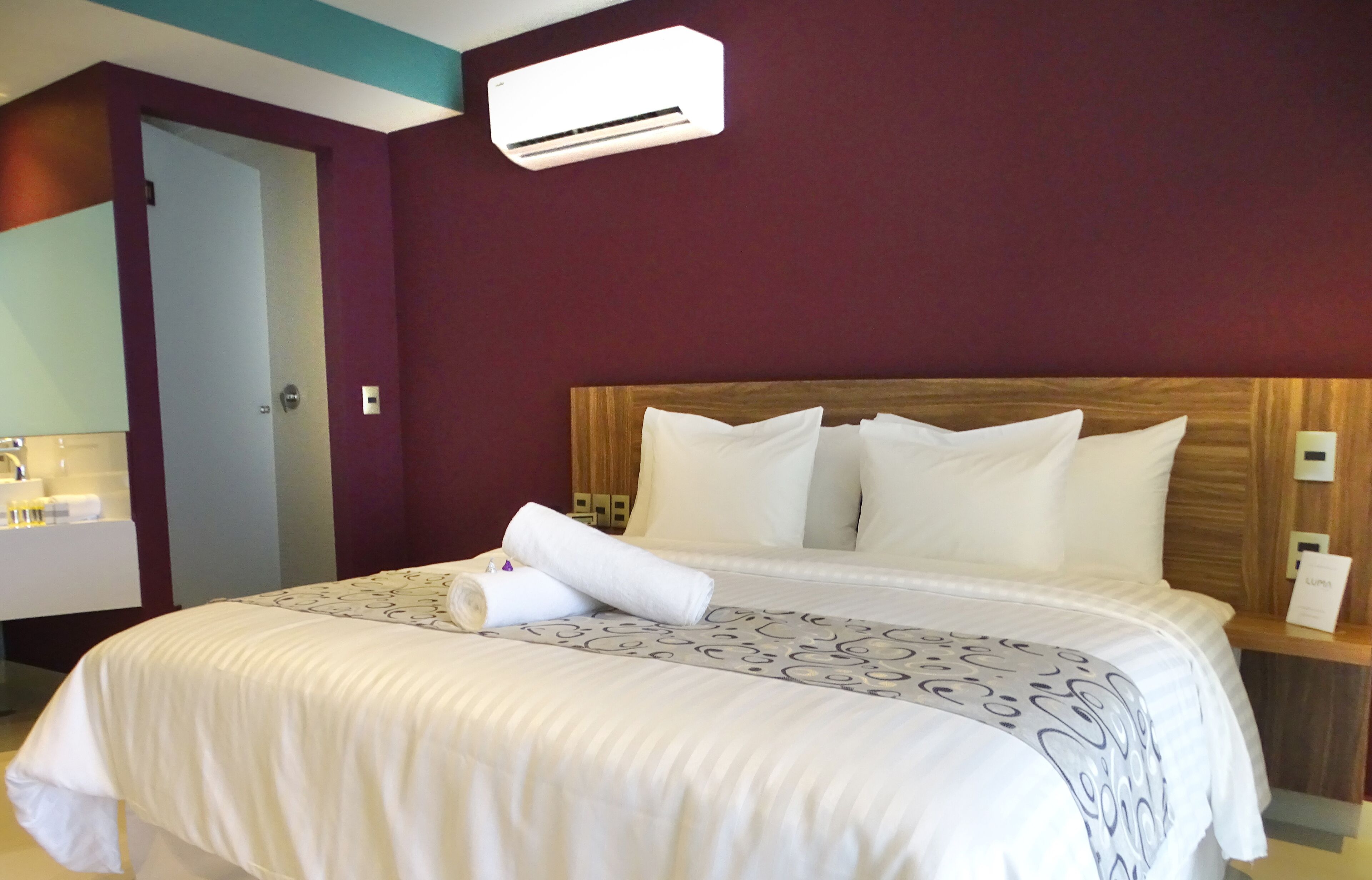 A hotel room with a large bed, a wooden headboard, white bedding, a bedside table with a lamp, and an air conditioning unit on the wall.