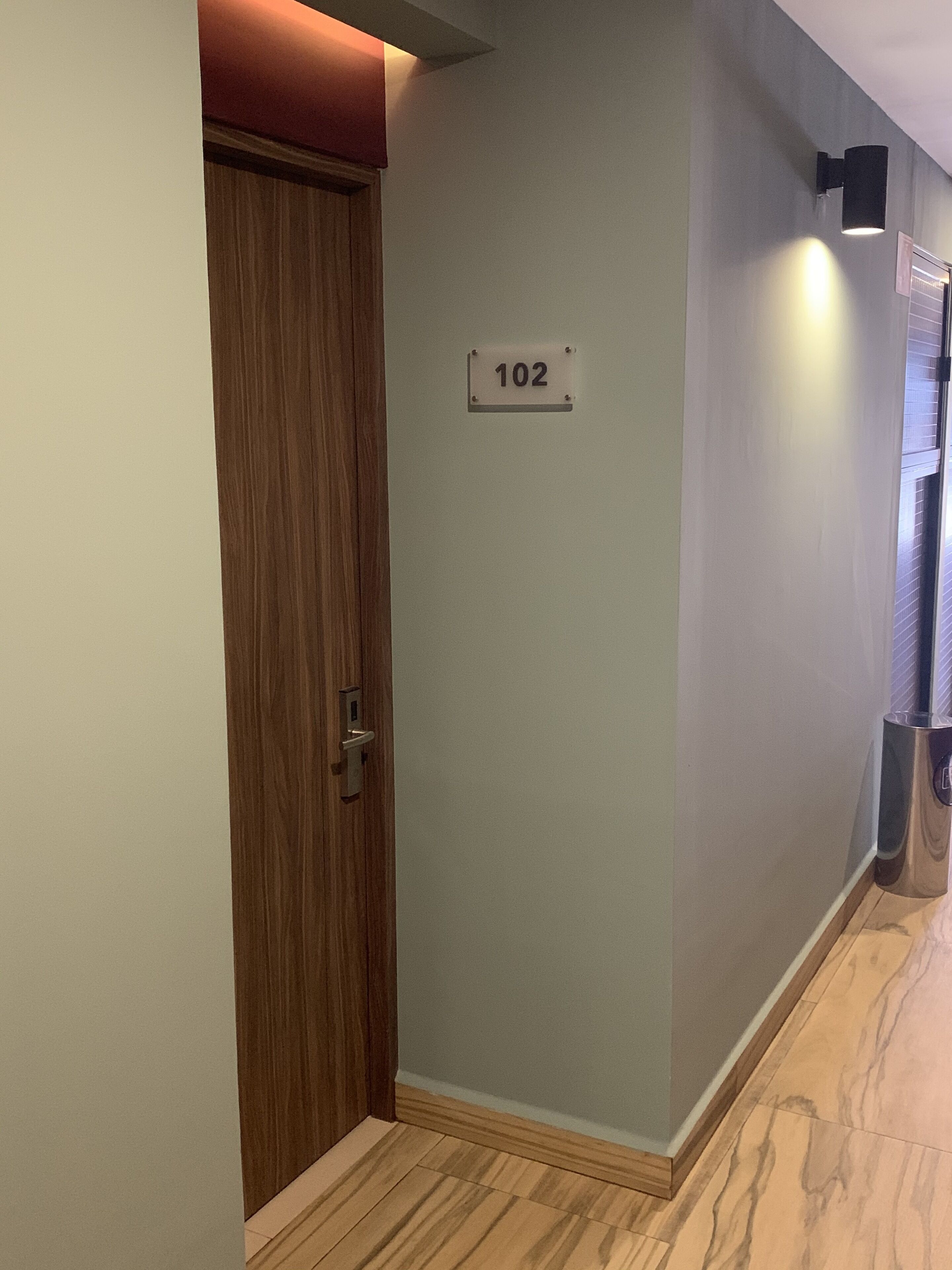 A hallway with a wooden door, a wall-mounted sign with the number 102, and a metallic trash can.