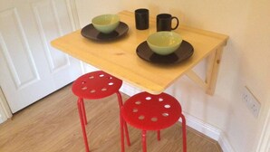 A small kitchen with a wooden table, two red stools, and three bowls and mugs on the table. - 1-bedroom Ground Floor Near Rugby School (Rugby)