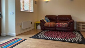 A cozy living room with a brown leather sofa, a patterned rug, and two wall-mounted lights. - 1-bedroom Ground Floor Near Rugby School (Rugby)
