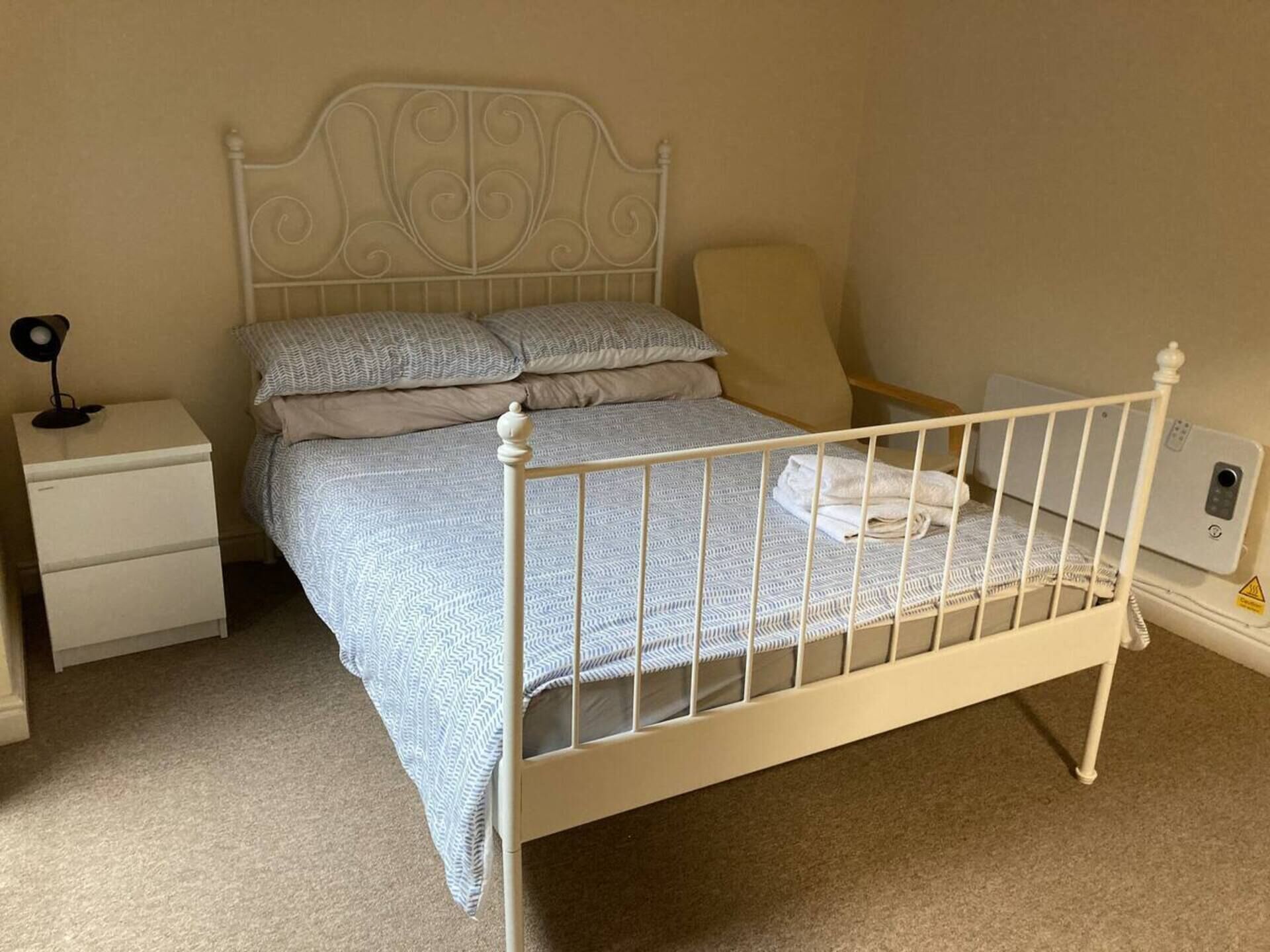 Una cama individual con estructura metálica blanca, cabecera con detalles ornamentales de hojas y una mesita de noche con lámpara.
