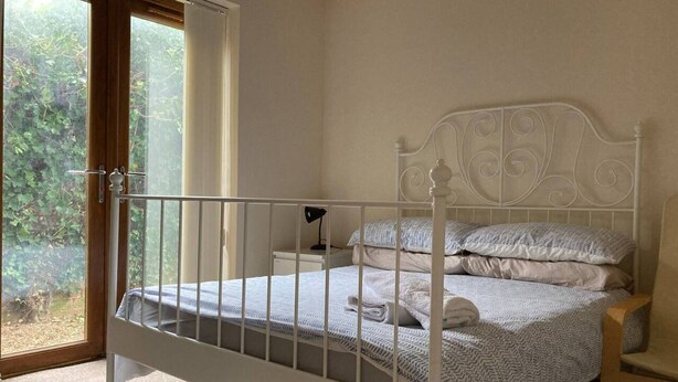A white metal bed with a headboard, a bedside table with a lamp, and a view of greenery through a sliding door. - 1-bedroom Ground Floor Near Rugby School (Rugby)