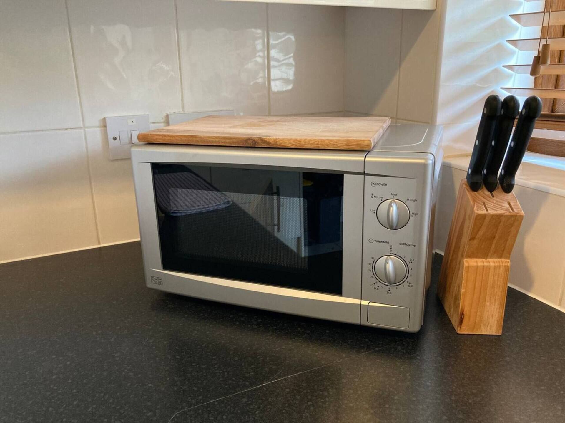 Un four à micro-ondes sur lequel est posée une planche à découper en bois, un bloc à couteaux contenant différents couteaux, et une prise électrique au mur.