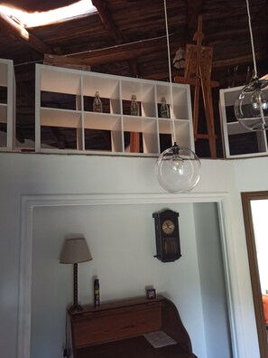 Una habitación con techo de madera, pared blanca, cómoda de madera, lámpara, reloj y repisa con botellas.