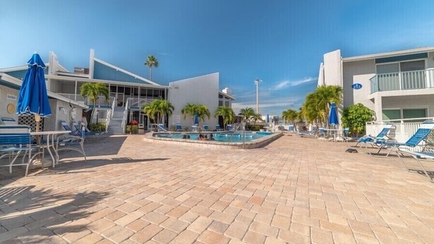 A pool area with lounge chairs and umbrellas, surrounded by palm trees and modern buildings.