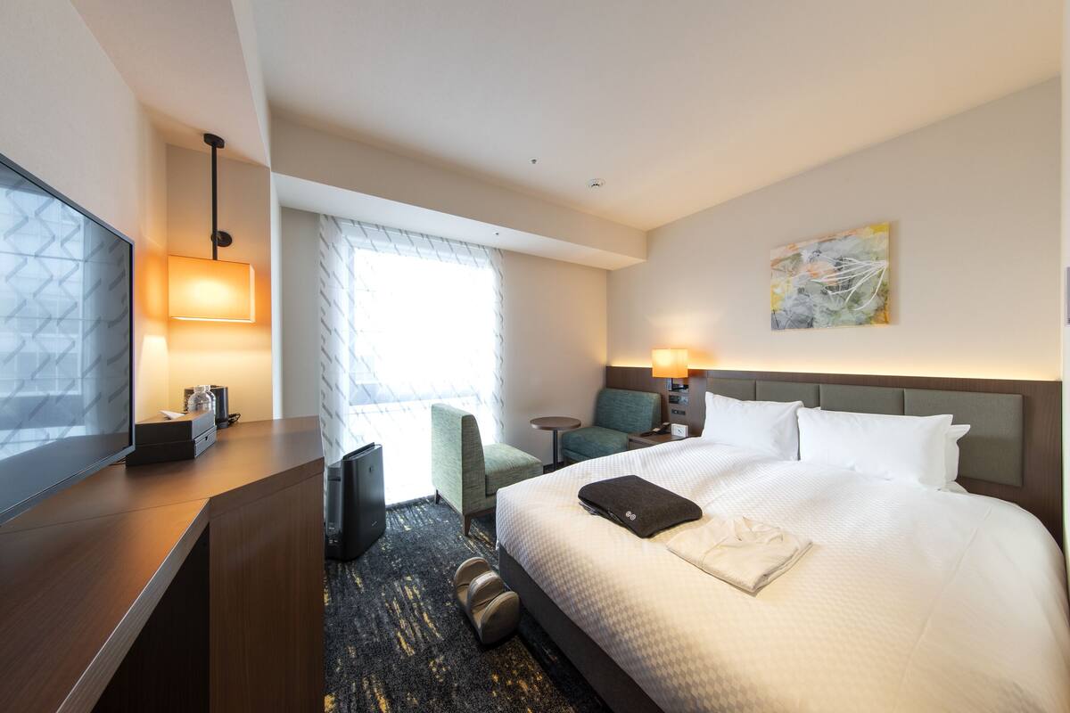 A hotel room with a large bed, a desk with a lamp, a chair, a small table, and a painting on the wall.
