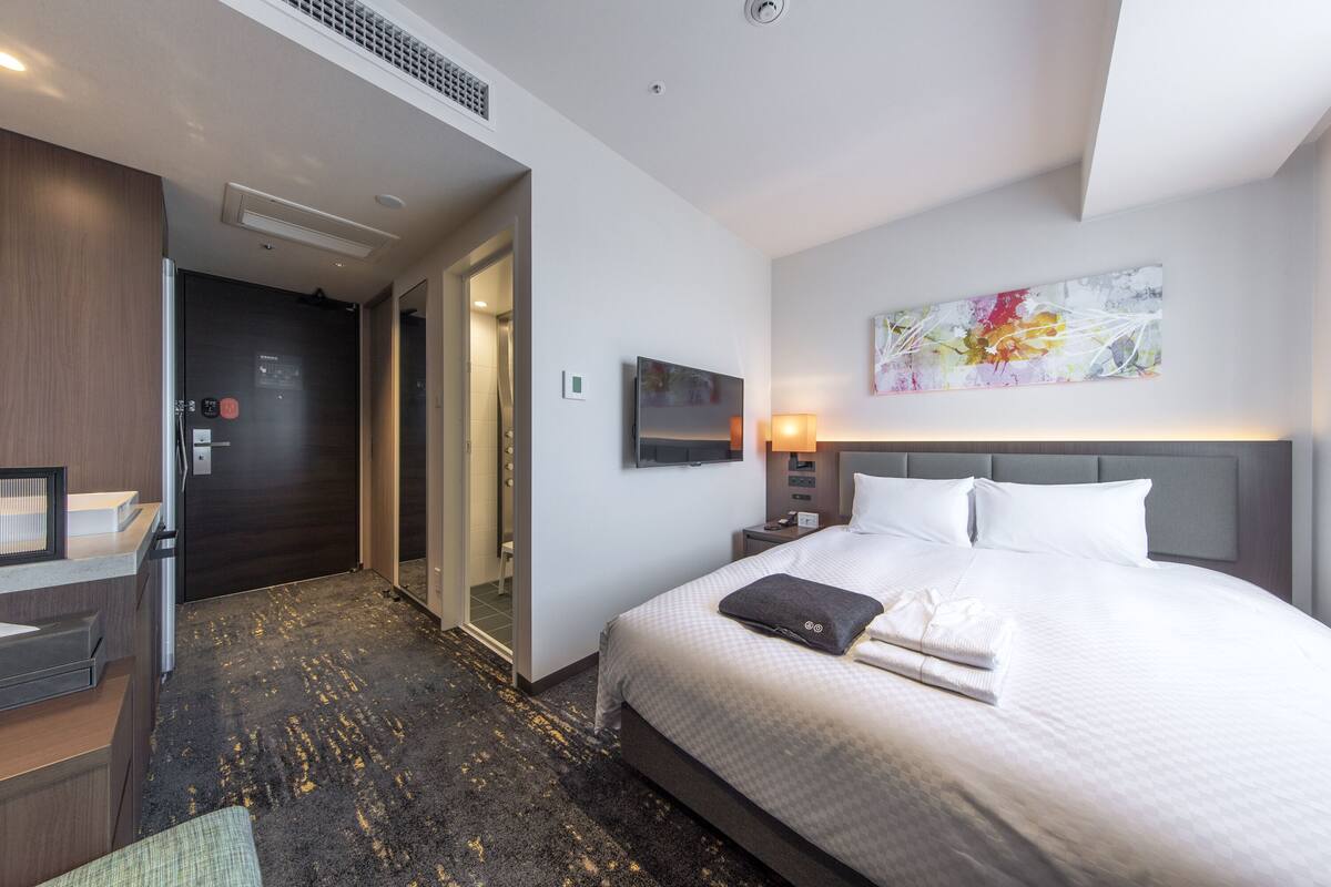 A hotel room with a bed, a desk, a flat-screen TV, a painting, and a bathroom with a shower and toilet.
