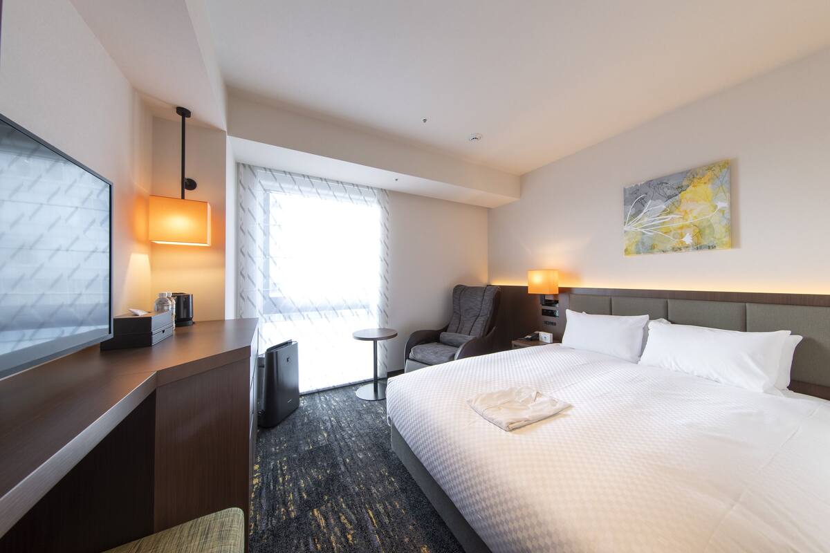 A hotel room with a large bed, a desk, a chair, a TV, and a window with blinds.