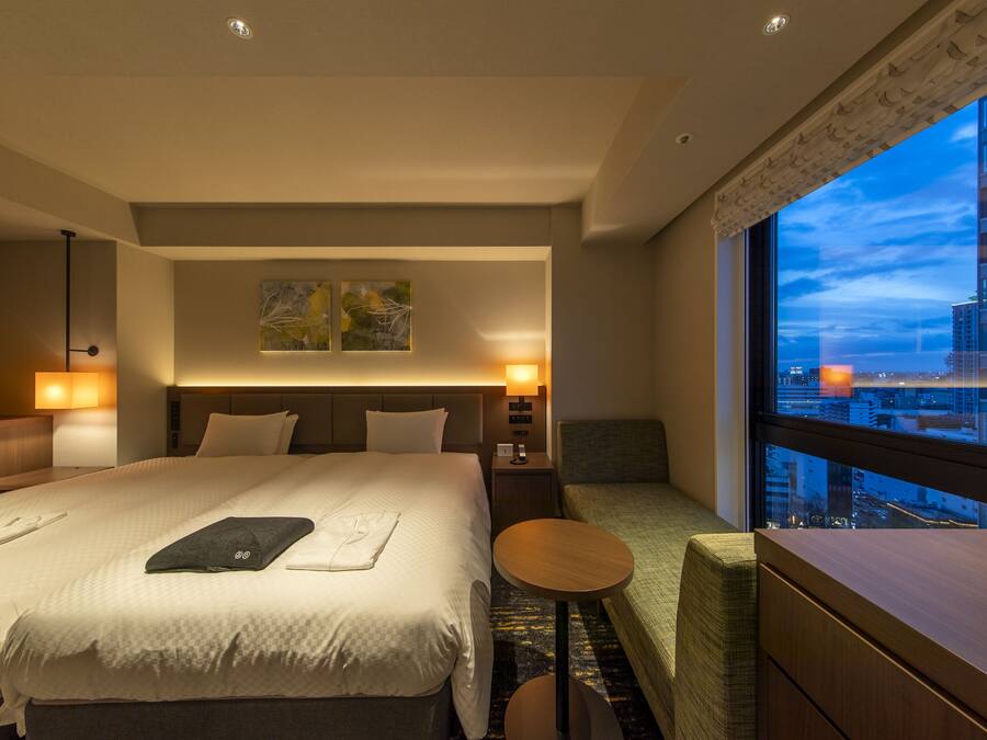 A hotel room with a large bed, two bedside tables, a small round table, and a view of the cityscape through a large window.