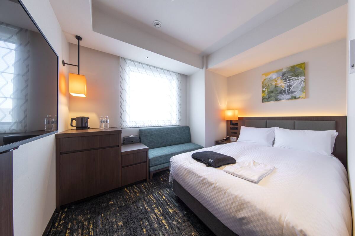 A hotel room with a bed, a desk, a chair, a window with curtains, a painting on the wall, and a lamp.