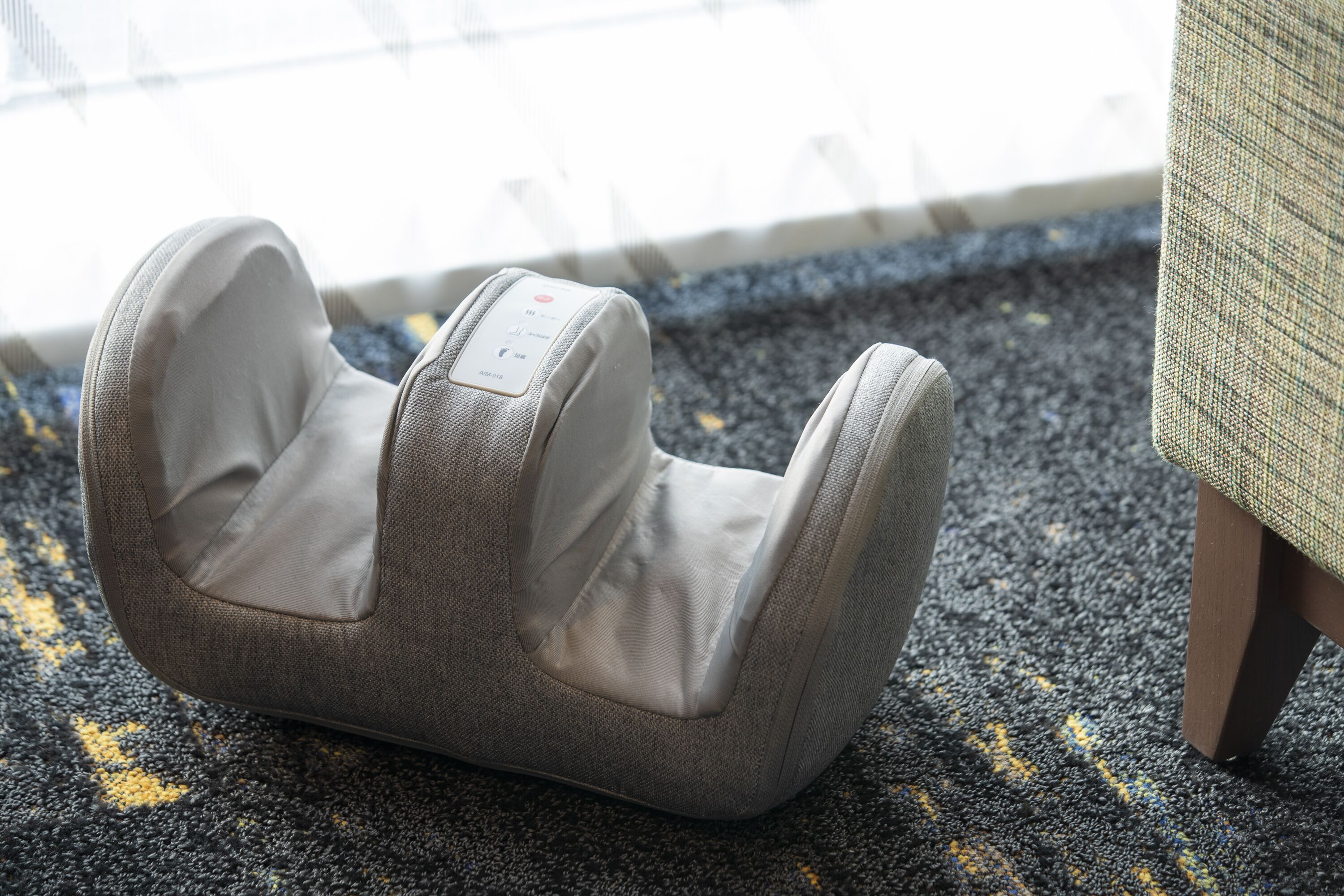 A massage chair with a control panel, positioned on a carpeted floor near a window.