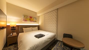 Habitación de hotel con una cama grande, una mesita de noche con lámpara, una pequeña mesa y un televisor montado en la pared.