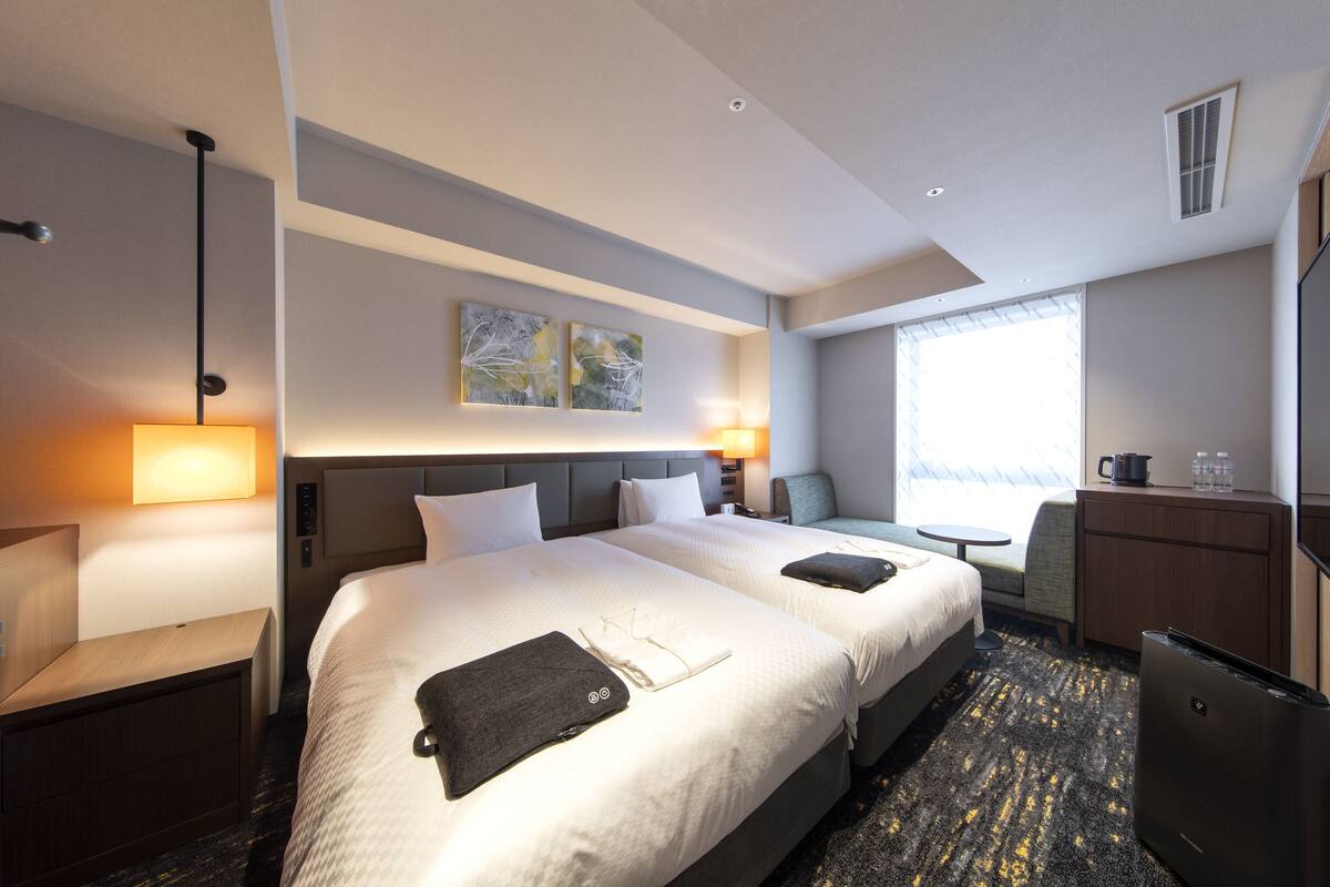 A hotel room with two beds, a desk, a chair, a small table, a refrigerator, and a window with a view.