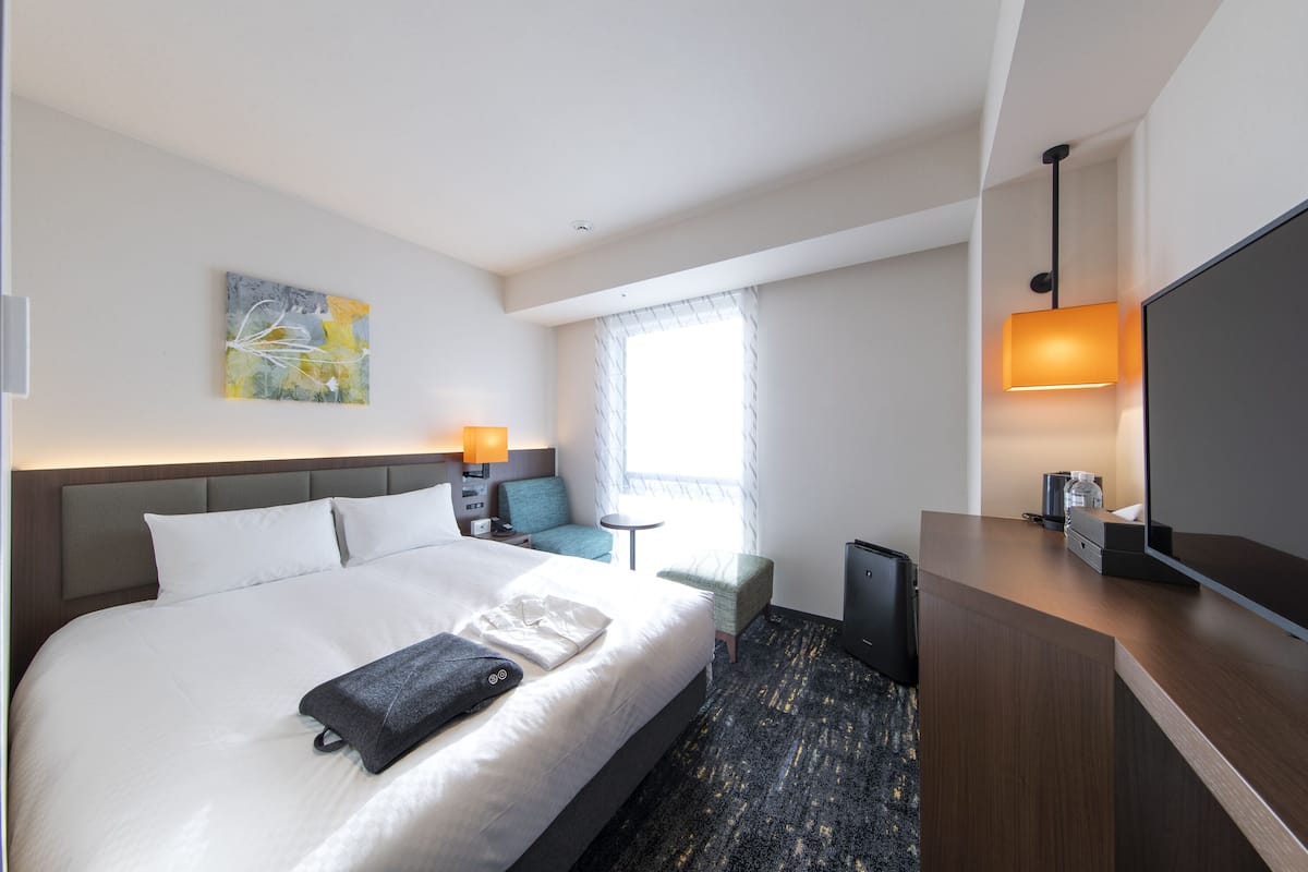 A modern hotel room with a large bed, a desk with a flat-screen TV, a small table, and a chair.