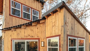 A wooden cabin with a deck, two wicker chairs with cushions, and a sliding glass door.