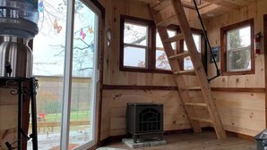 A wooden cabin interior with a fireplace, large windows, and a ladder.