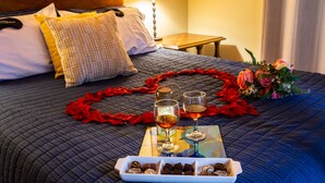 Een net opgemaakt bed met een gewatteerde deken, kussens, een nachtkastje met een lamp, een schaal met chocolade, twee glazen wijn en een bloemenboeket.