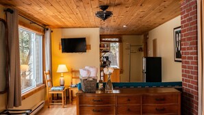 Een gezellige kamer met een houten plafond, een aan de muur bevestigde televisie, een dressoir met lades, een stoel, een lamp en een raam met gordijnen.