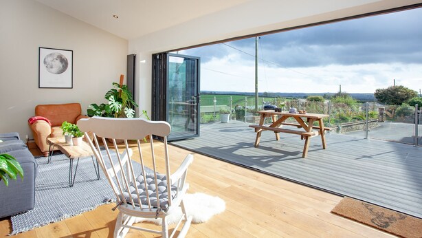 A modern living room with a sofa, a rocking chair, a dining table, and a large sliding glass door leading to an outdoor deck. - 3 bedroom accommodation in Blunts, near Saltash (Blunts, near Saltash)