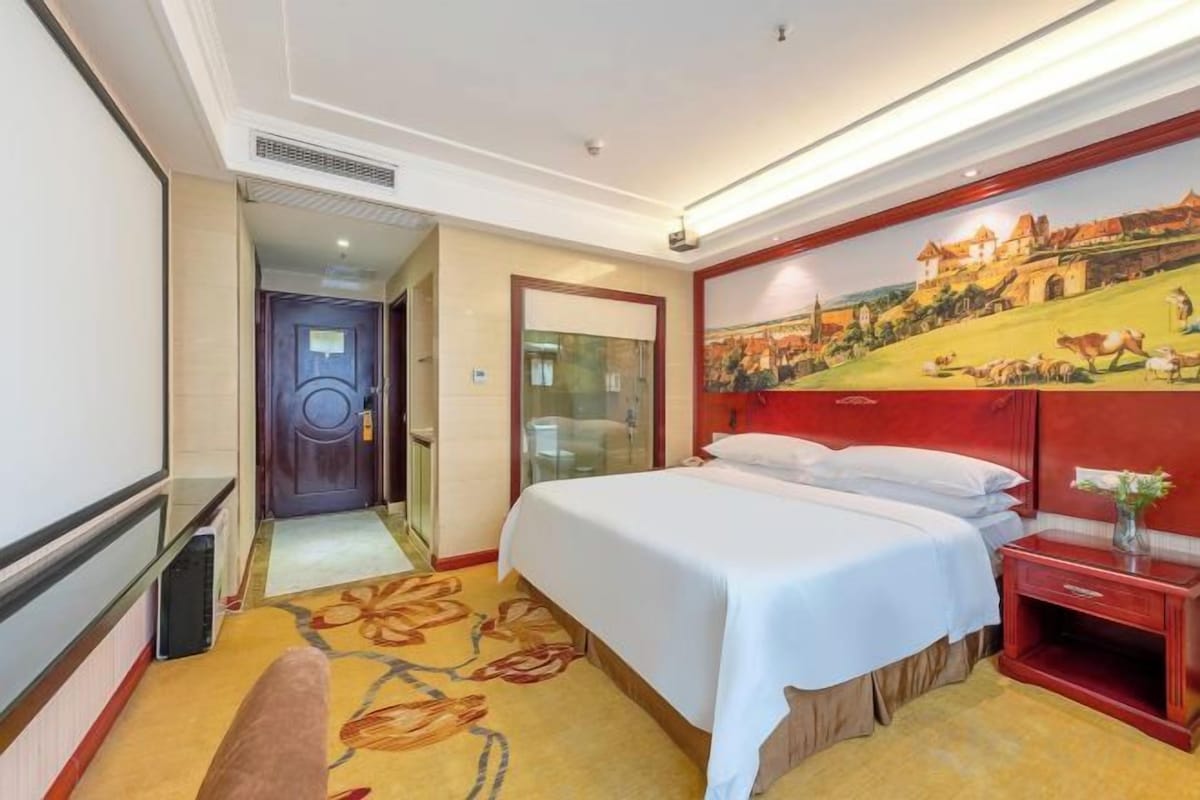 Quarto de hotel com cama, escrivaninha, cadeira, pintura na parede e porta que dá para outro cômodo.