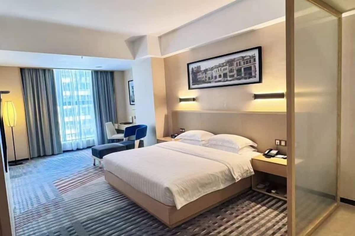 A hotel room with a bed, bedside table, lamp, chair, and a large window with curtains.