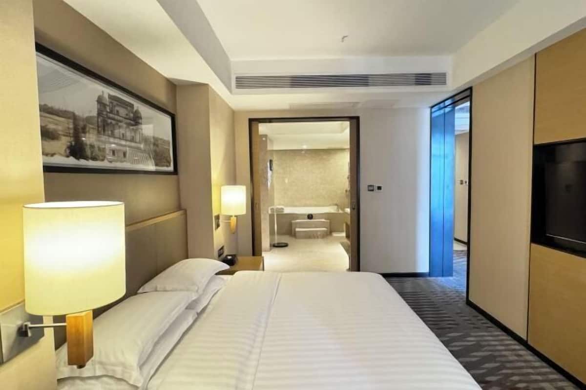 A hotel room with a bed, a nightstand, a lamp, a TV, a framed picture on the wall, and a bathroom visible through an open door.