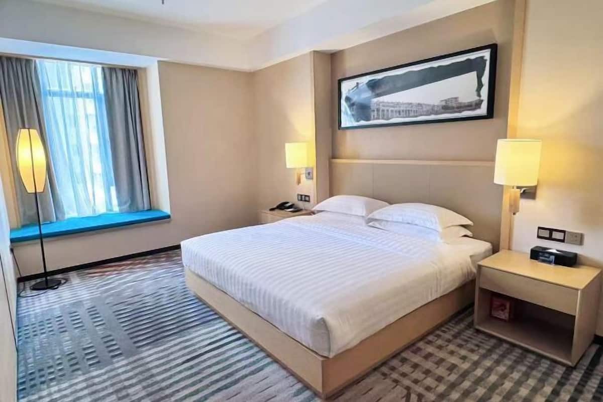 A hotel room with a bed, bedside lamps, a window with curtains, a wall-mounted picture, and a bedside table.