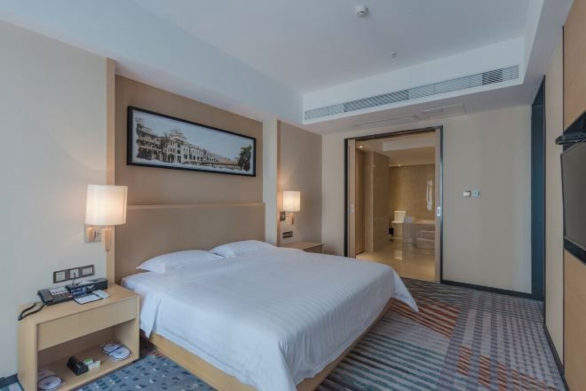 A hotel room with a bed, bedside tables, lamps, a framed picture on the wall, and a door leading to a bathroom.