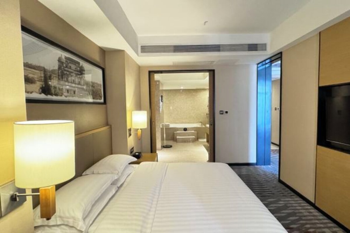 A hotel room with a bed, a nightstand, a lamp, a TV, a framed picture on the wall, and a bathroom visible through an open door.