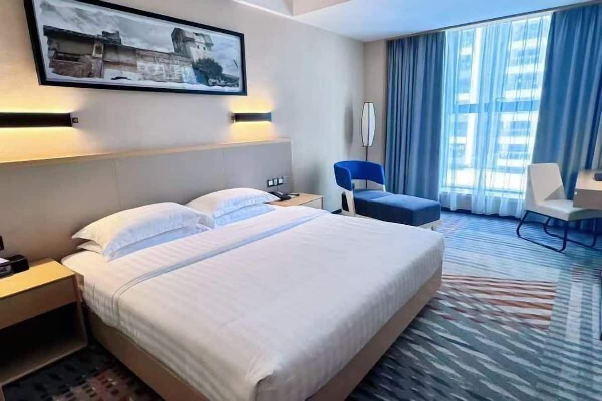 A hotel room with a large bed, a desk, a chair, a desk lamp, a painting on the wall, and a view of the city through the window.