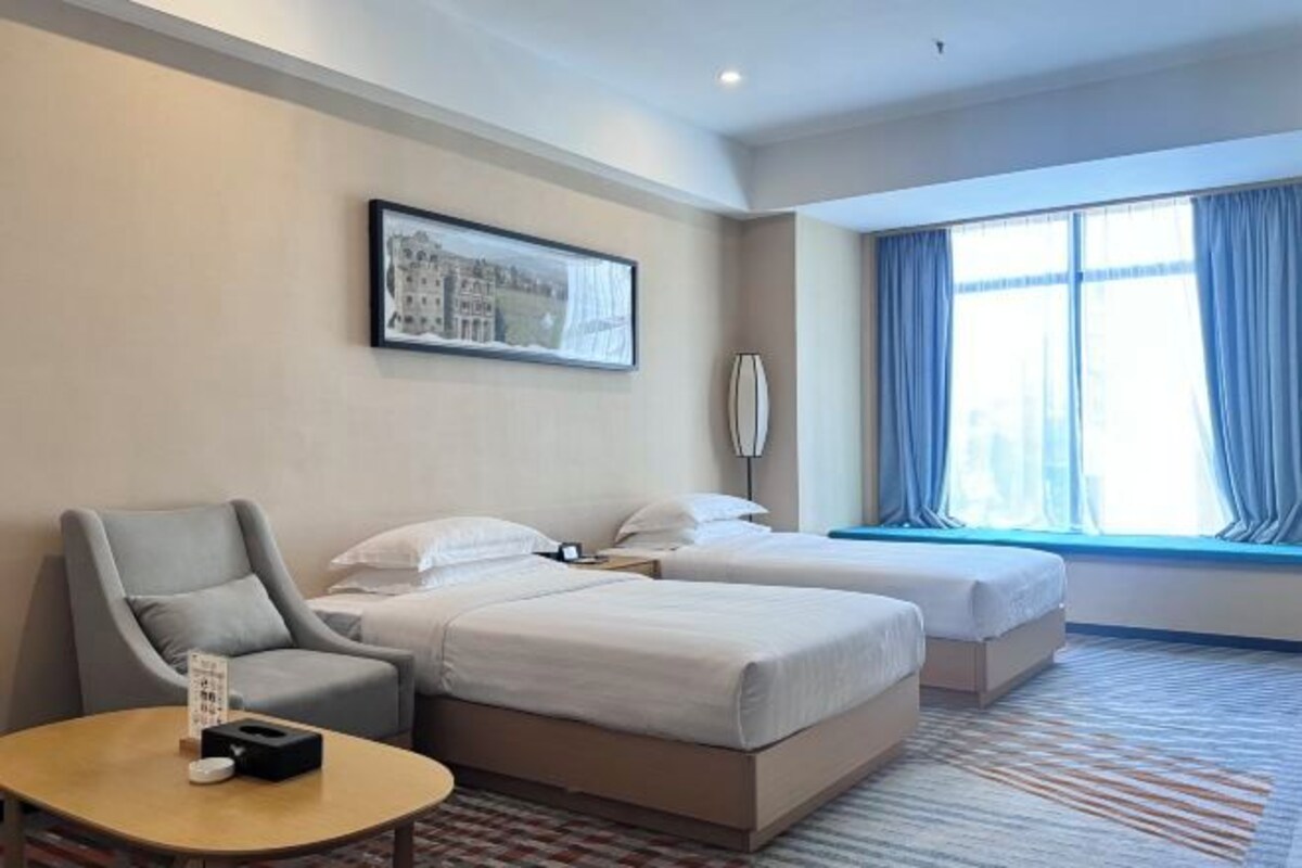 A hotel room with two beds, a chair, a small table, a painting on the wall, and a large window with curtains.