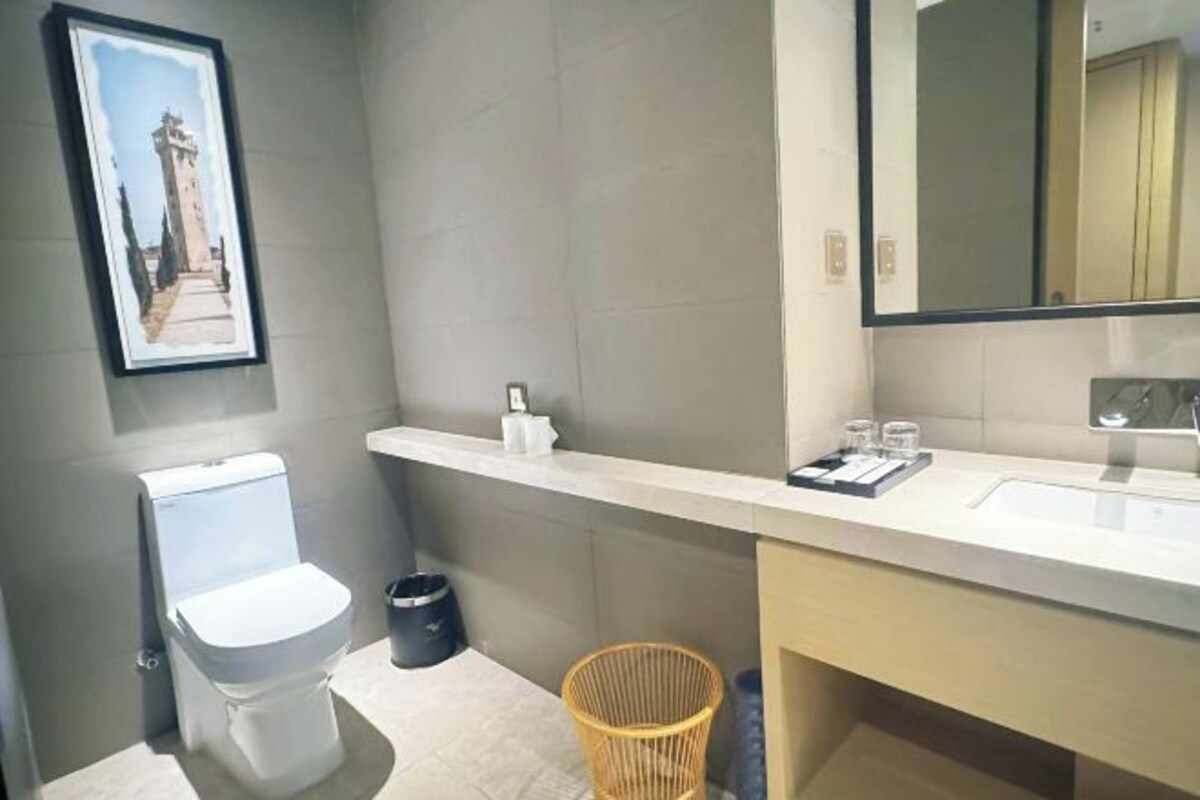 A bathroom with a toilet, a trash bin, a wicker basket, a shelf with toiletries, and a framed picture on the wall.