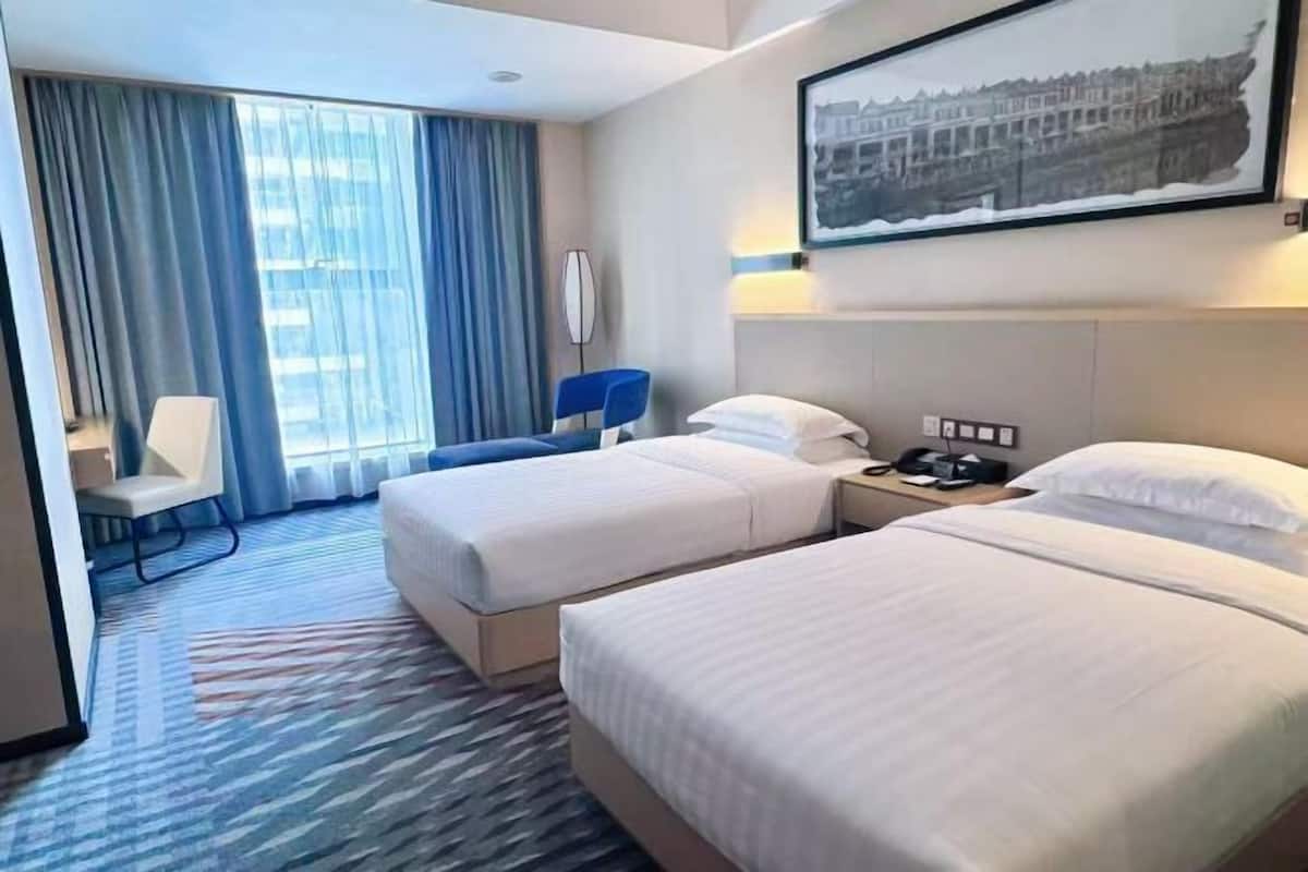 A hotel room with two beds, a desk, a chair, a large window with curtains, and a framed picture on the wall.