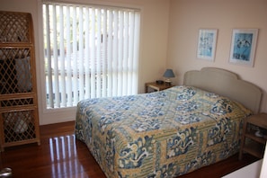A bedroom with a bed, a nightstand, a lamp, a window with blinds, and a wooden shelf. - Seachange (Forster)
