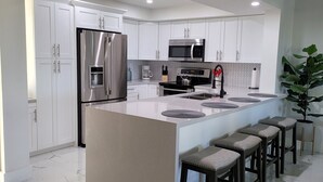 A modern kitchen with white cabinets, stainless steel appliances, a marble countertop, and a central island with bar stools. - Coastal Escape ● Steps to Beach ● Sunset Views (Fort Myers Beach)