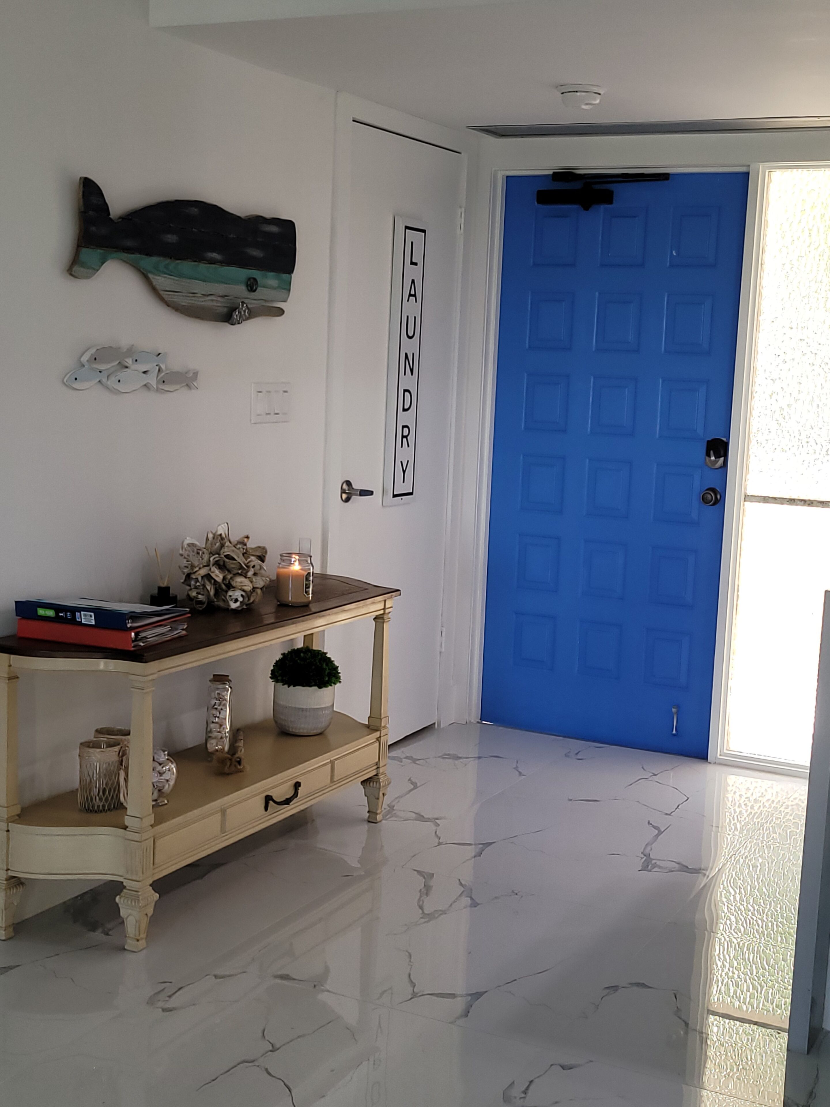 Un couloir avec une porte bleue portant l’inscription « buanderie », une console en bois ornée d’objets décoratifs et une œuvre d’art murale représentant des poissons.