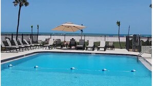 A clear blue pool with lounge chairs and umbrellas, surrounded by a fence and palm trees, with a view of the ocean. - Coastal Escape ● Steps to Beach ● Sunset Views (Fort Myers Beach)