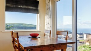 Un comedor con una mesa y sillas de madera, un cuenco rojo y vistas al mar y las colinas a través de amplias ventanas.