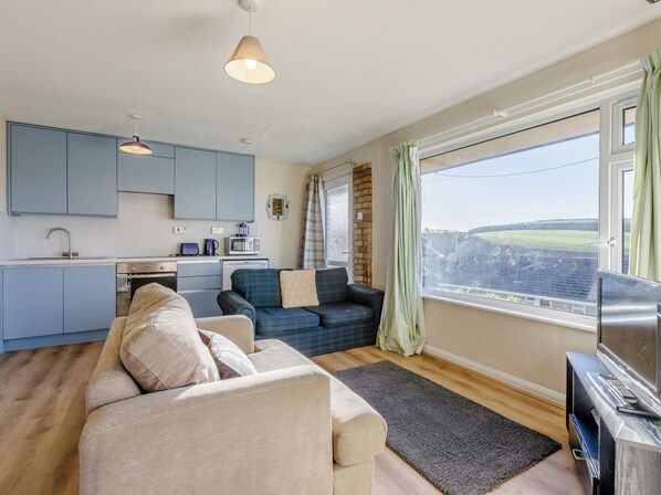 A modern kitchen with blue cabinets, a dining area with a sofa and a TV, and a large window offering a view of the countryside.