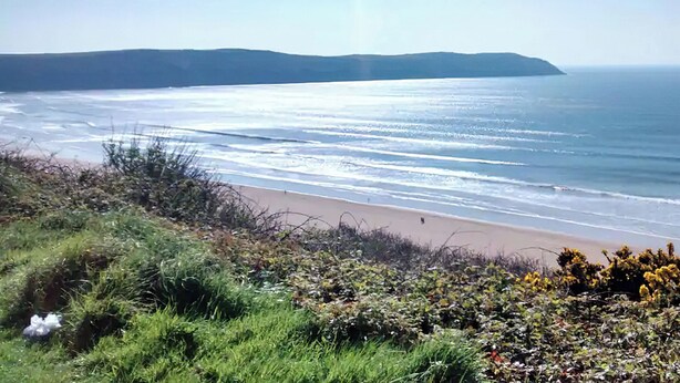 Paisaje costero con playa, primer plano de hierba y colinas al fondo.