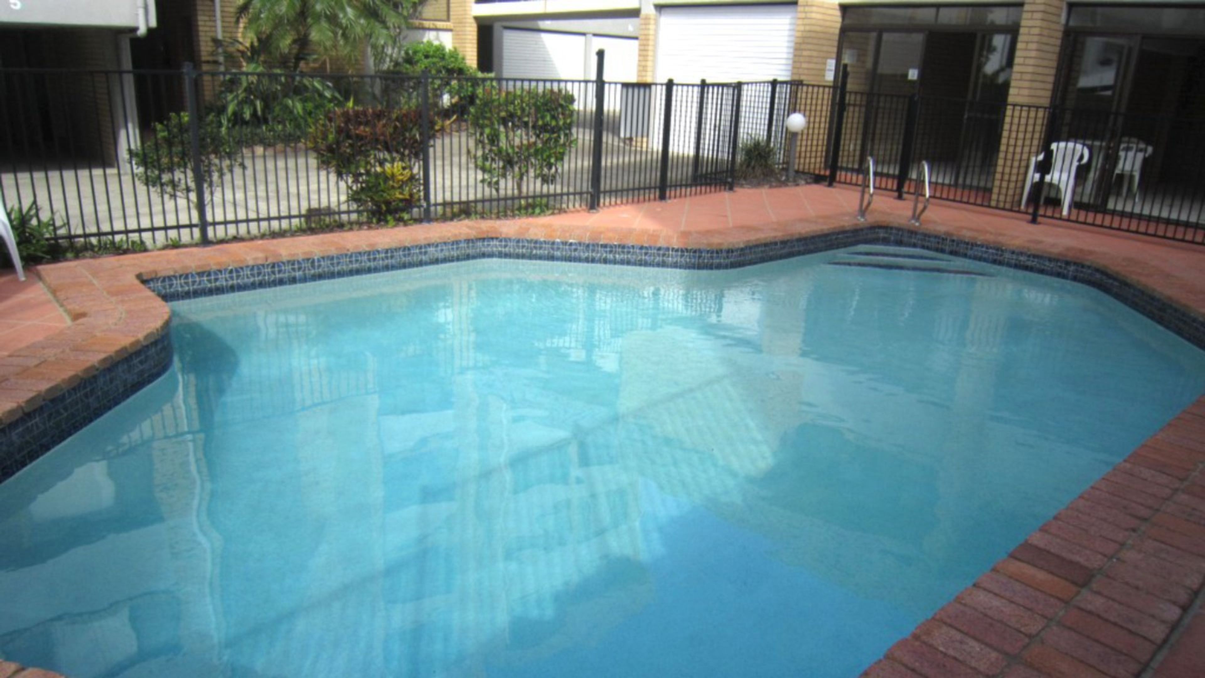 Une piscine rectangulaire dont les bords sont recouverts de carreaux, entourée d’une bordure en briques et d’une clôture en métal.
