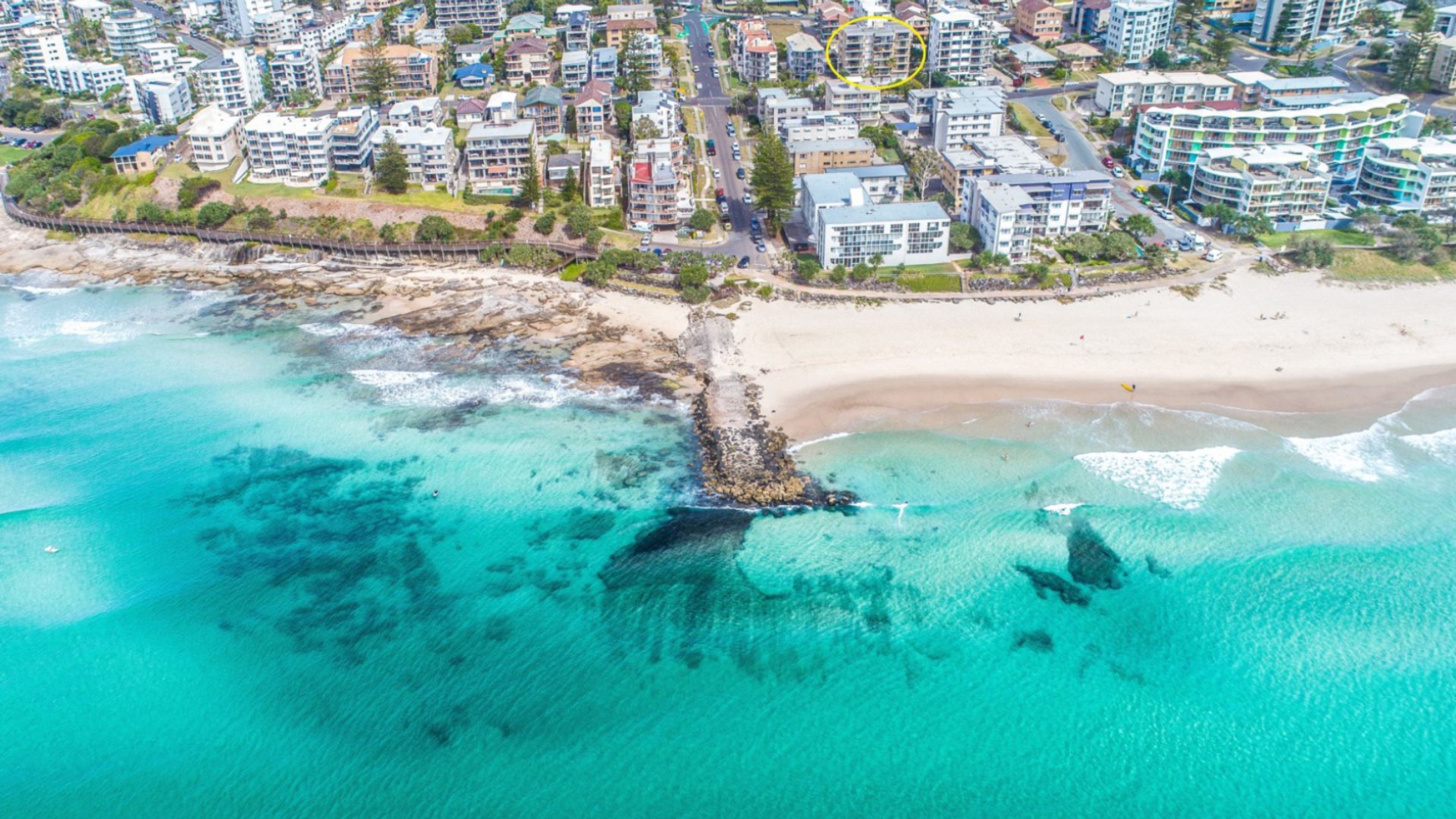 Vue aérienne d’une zone côtière avec une plage, des eaux turquoise et limpides, et une zone résidentielle dense avec de nombreux immeubles.