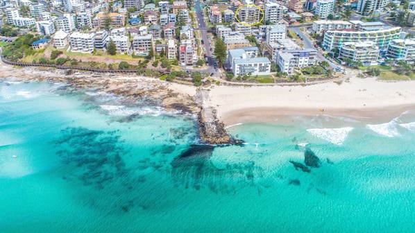 Vista aérea de una zona costera con playa, aguas turquesas cristalinas y una densa área residencial con edificios de varios pisos.