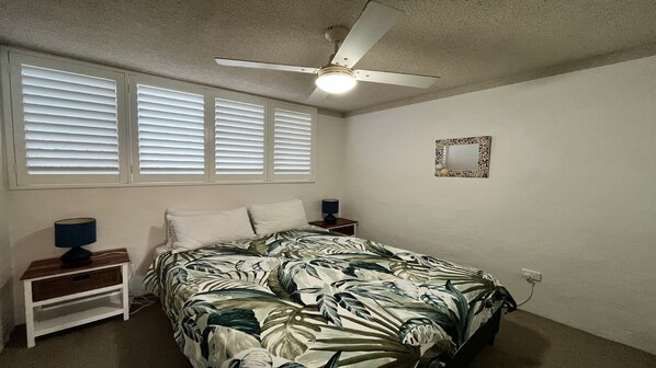 A bedroom with a bed, two bedside tables, a ceiling fan, a window with blinds, and a wall-mounted mirror.