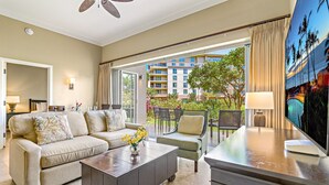 A hotel room with a sofa, coffee table, and chairs, a balcony with a view of buildings and greenery, and a painting on the wall.