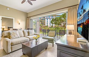 A hotel room with a sofa, coffee table, and chairs, a balcony with a view of buildings and greenery, and a painting on the wall. - Maui Resort Rentals: Honua Kai Hokulani 144 – Large Ground Floor 1BR, Spacious Lawn Area + Easy Pools/Beach Access (Lahaina)