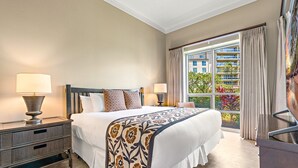 A hotel room with a large bed, two bedside lamps, a nightstand, a dresser, and a view of a building and greenery through the window.
